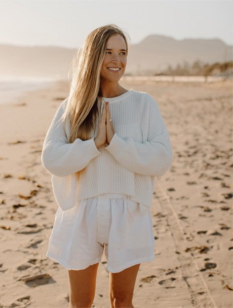 Jordan (The Balanced Blonde) wearing a white sweater and white shorts on a beach at sunset.